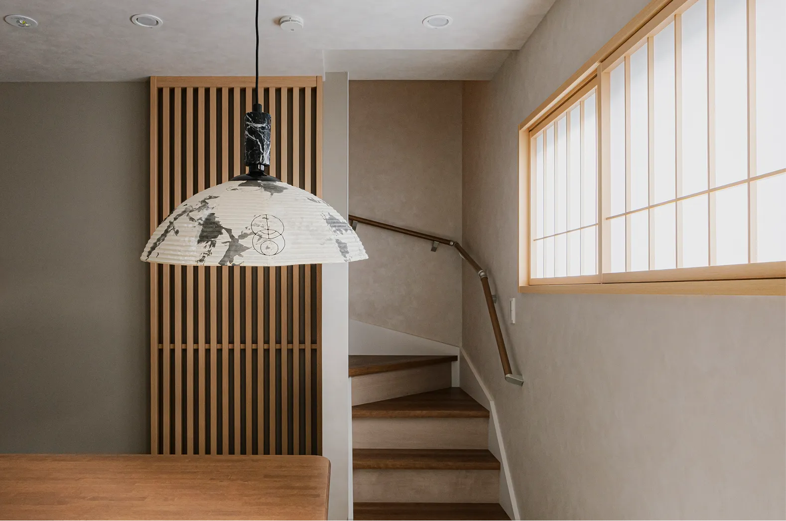 木目と白を基調にした温かみのある室内。天井から吊り下げられた照明と、奥には階段のある和風な空間。