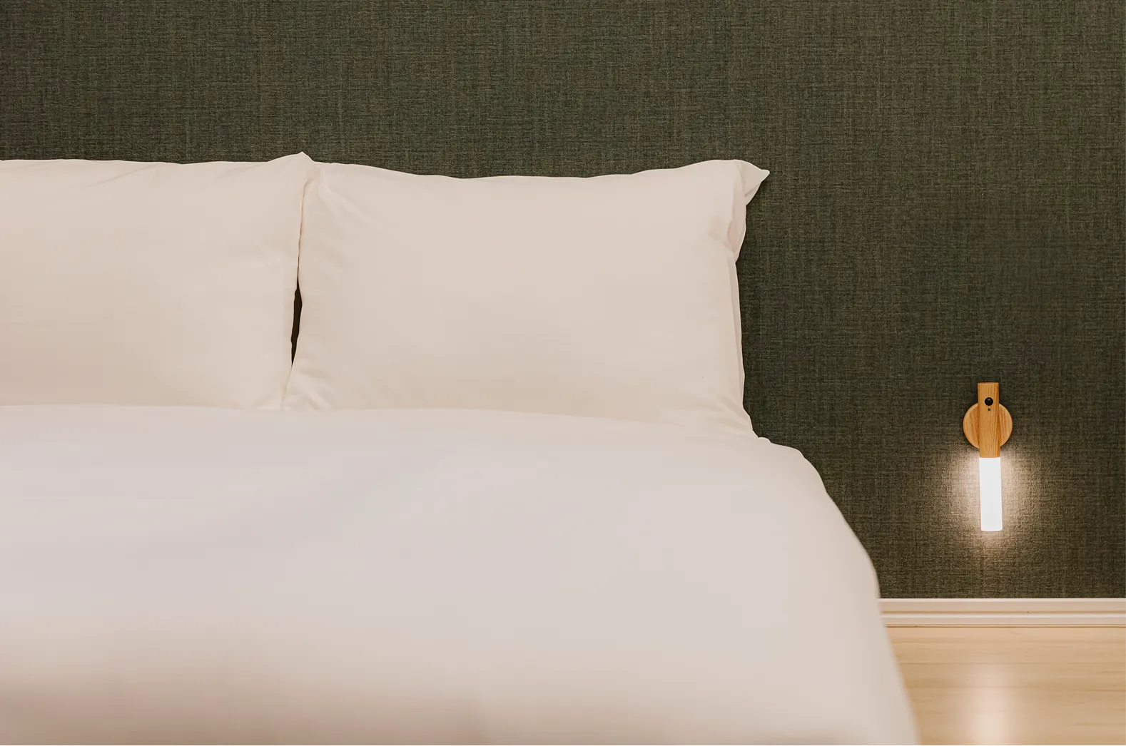 緑のアクセントウォールが印象的な寝室。白いベッドに、足元にはライトが設置されている。