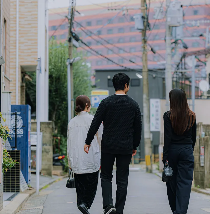 電柱や電線が張り巡らされた路地を歩く3人の男女の後ろ姿。