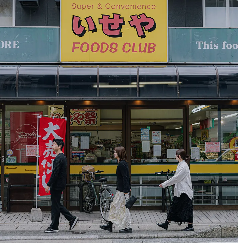 黄色い看板のスーパー「いせや」の前を横切る3人の男女。