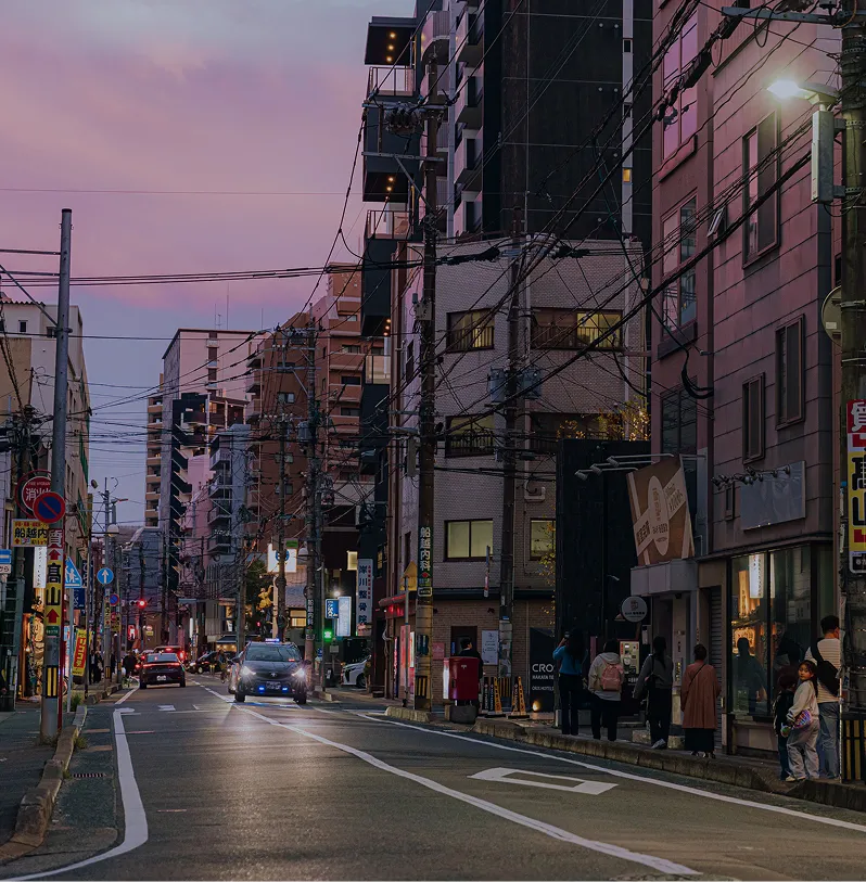 紫色の夕焼け空の下、車や歩行者が行き交う街角の風景。