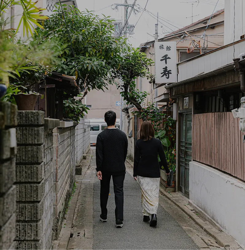 細い路地を歩き去る、黒い服を着た男女の後ろ姿。