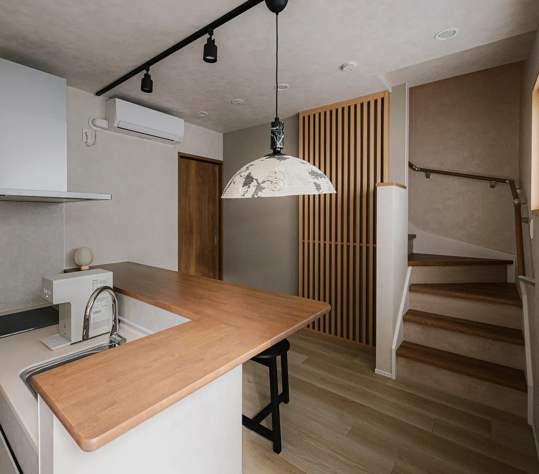 L字型の木製カウンターが特徴的なキッチン。頭上には和紙のような質感のペンダントライトが灯り、奥には格子状の引き戸と上階へ続く木の階段が見える、落ち着いた色調のジャパンディスタイルの室内。