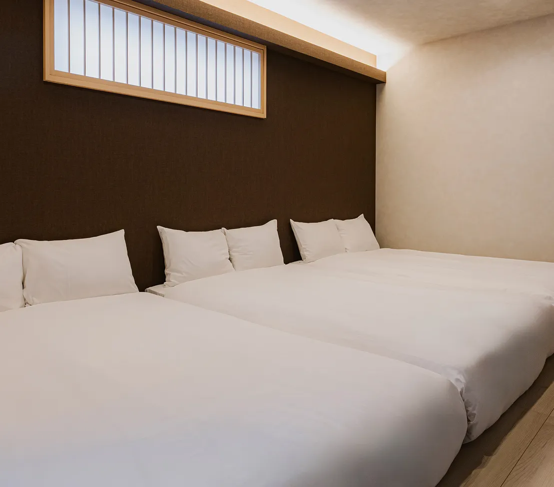 ダークブラウンの壁に障子風の窓が配置された、モダンで落ち着いた雰囲気の寝室。清潔感のある真っ白なリネンで整えられたベッドが3台並び、天井からの柔らかな間接照明が心地よい眠りを誘うジャパンディな空間。